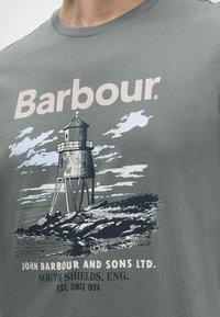 Πράσινο πουκάμισο με το λογότυπο Barbour και γραφικό φάρου δίπλα σε κείμενο για την εταιρεία John Barbour and Sons Ltd., South Shields, Αγγλία.