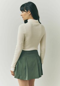 Cremefarvet ribbestrikket turtleneck sweater og grøn plisseret nederdel, der fremhæver en tætsiddende silhuet og glatte teksturer.