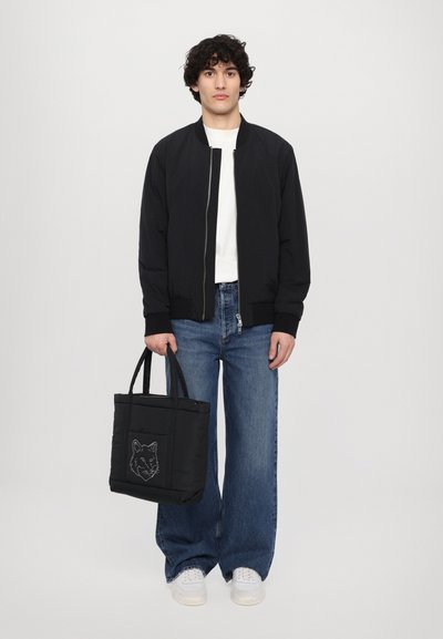 Black bomber jacket, white t-shirt, blue wide-leg jeans, and a black tote bag with a wolf design. White sneakers complete the outfit.