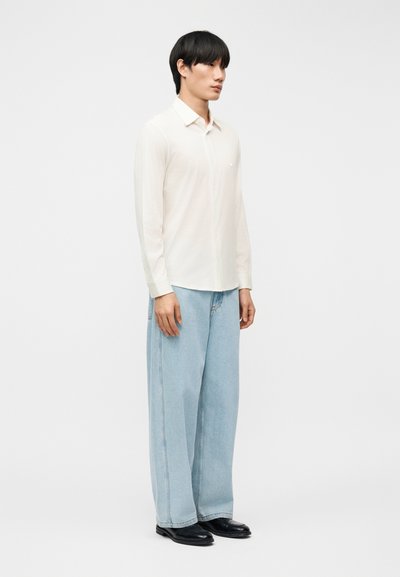 Νέος άντρας που στέκεται φορώντας λευκό πουκάμισο με μακριά μανίκια, ανοιχτό μπλε τζιν πλατύ στο κάτω μέρος και μαύρα παπούτσια, σε απλό λευκό φόντο.
