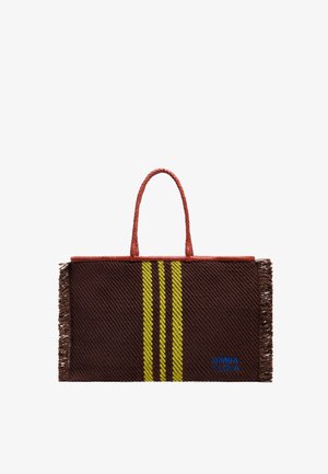 Woven brown tote bag with yellow stripes, red braided handles, and fringe detail. Features blue branding at the bottom.
