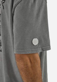 Κοντινό πλάνο του μπράτσου ενός ατόμου που φορά γκρι κοντομάνικο πουκάμισο με μικρό στρογγυλό λευκό λογότυπο κοντά στο τελείωμα του μανικιού.