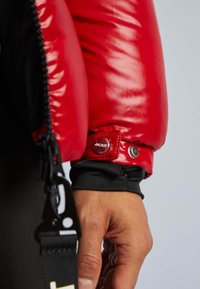 Giacca imbottita rossa con finitura lucida, dotata di polsini aderenti, bottone a pressione con il marchio "JACKIT" e dettagli in tessuto nero.
