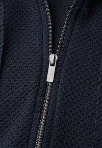 Gros plan sur une fermeture éclair argentée sur un tissu texturé foncé, partiellement fermée avec des dents métalliques visibles et une languette de tirage centrée verticalement.