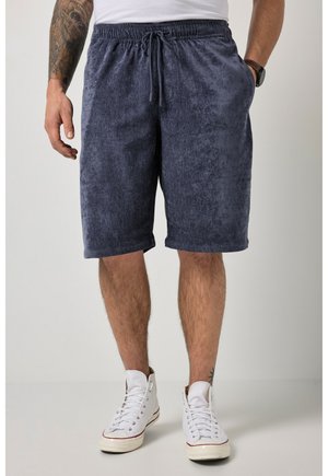 Uomo che indossa pantaloncini in spugna blu navy con vita elastica e coulisse, sneakers bianche alte e camicia bianca, in piedi davanti a uno sfondo semplice.