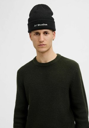 Zwarte gebreide beanie met "JJ Studios" geborduurd in wit. Het model draagt een gestructureerde donkergroene sweater met een ronde hals.
