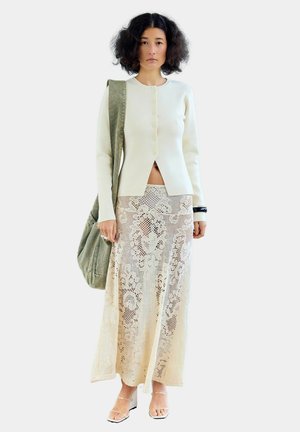 Femme aux cheveux bouclés porte un pull crème boutonné, une jupe maxi en dentelle beige, des talons à brides transparentes, et porte un sac à bandoulière vert olive.