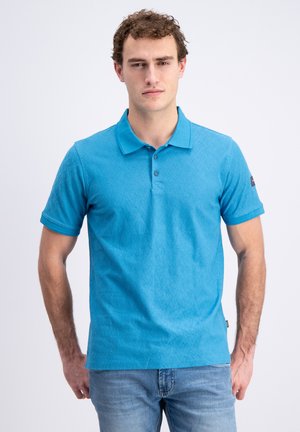 Uomo con capelli ricci che indossa una polo blu a fantasia e jeans azzurri, in piedi su uno sfondo chiaro uniforme.