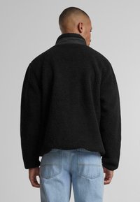 Jachetă din fleece neagră cu guler înalt, material texturat și croială lejeră. Asortată cu blugi decolorati de culoare albastră deschisă, evidențiind un stil casual.