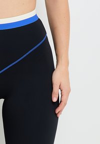 Leggings noirs taille haute avec une ceinture bleu et blanche, présentant une ligne d'accent bleu et une texture lisse et extensible.