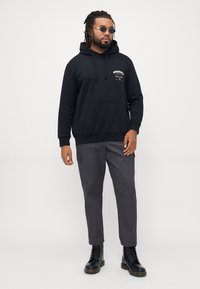 Black hoodie with front pocket and logo, grey cargo pants with side pockets, and black lace-up boots. Features sunglasses and braided hair.