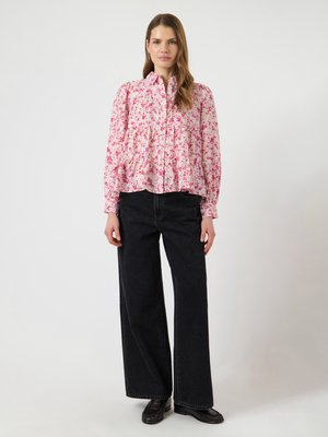 Kvinne iført en langermet rosa blomstret bluse og svart, vidbukset, høytlivs jeans med svarte loafers, stående foran en ensfarget bakgrunn.