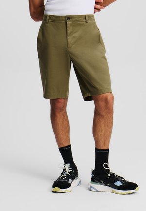 Shorts verde oliva con taglio dritto e tasche laterali, abbinati a sneakers bianco e nere con dettagli azzurri e verdi.