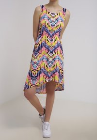 Femme portant une robe sans manches à motifs géométriques multicolores avec un ourlet asymétrique et des baskets blanches, se tenant les mains derrière le dos.