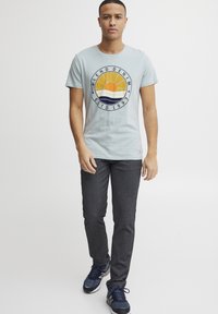 Lyseblå bomull T-skjorte med sirkulært grafisk motiv av en sol og bølger, paret med mørkegrå bukser og marineblå sneakers.