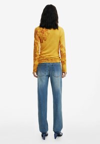 Langärmliges gelbes Oberteil mit floralen Mustern, kombiniert mit ausgewaschenen blauen weiten Jeans. Modell trägt marineblaue Absatzsandalen, von hinten betrachtet.