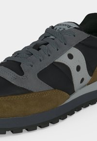 Κοντινή λήψη ενός αθλητικού παπουτσιού Saucony Jazz με μαύρες, γκρίζες, λευκές και καφέ σουέτ επιφάνειες και γκρι κορδόνια σε μια ανάγλυφη σόλα.