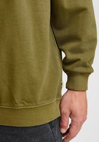 Oliwkowy sweatshirt z ribbowanym dołem i mankietami, o miękkiej fakturze i detalach wzdłuż bocznych szwów, z widoczną metką marki blisko dołu.
