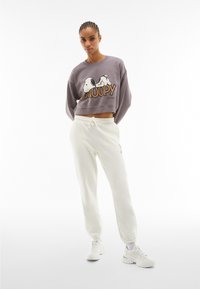 Sweatshirt gris court avec un motif Snoopy et un pantalon de survêtement blanc, tous deux en tissu doux. Le modèle porte des baskets blanches et se tient devant un fond uni.