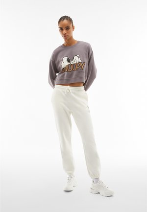 Felpa grigia corta con grafica di Snoopy e pantaloni della tuta bianchi, entrambi realizzati in tessuto morbido. Il modello indossa sneaker bianche e si trova di fronte a uno sfondo neutro.