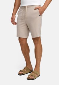 Hombre vestido con pantalones cortos beige de cordón y sandalias marrones, de pie con una mano en el bolsillo frente a un fondo blanco.