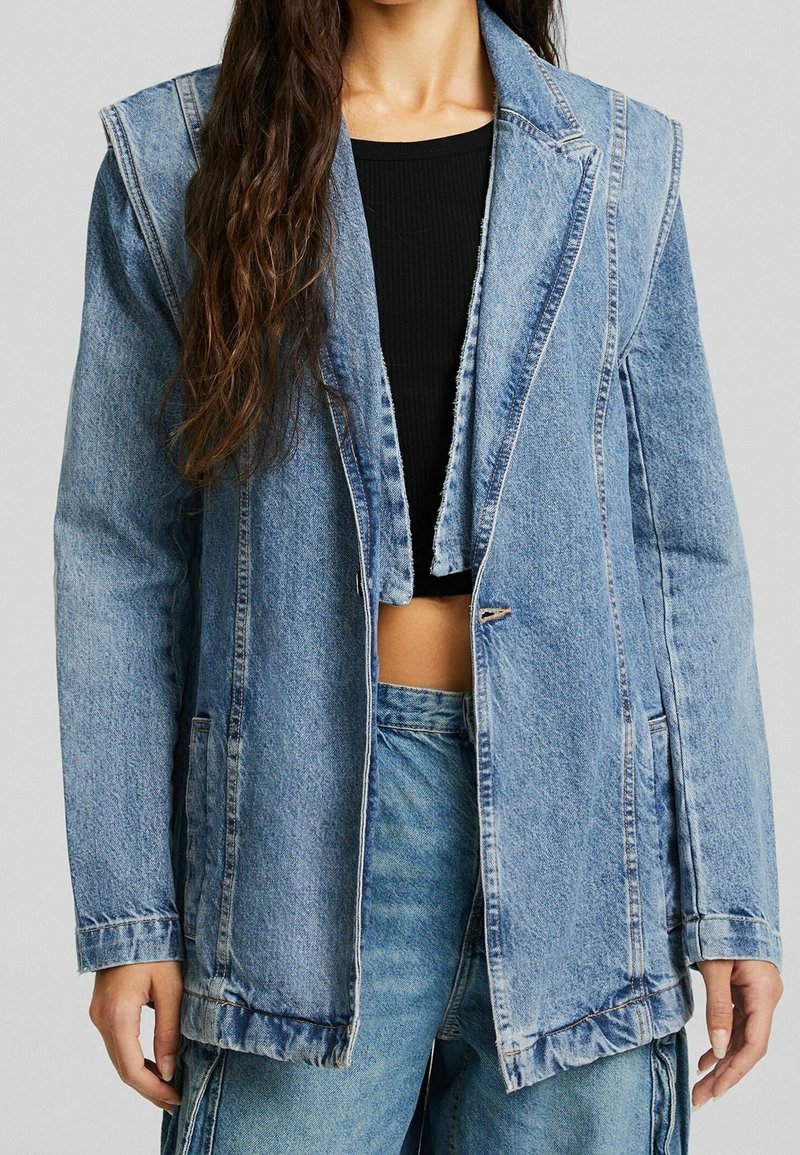 Světle modrá oversized džínová bunda s nezpracovanými okraji, klopami, dvěma předními kapsami a jedním knoflíkovým uzávěrem. Nanesena přes černý top.