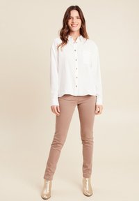 Witte overhemd met knoopsluiting en een borstzak, gecombineerd met lichtbruine skinny pants en glanzende gouden enkellaarsjes. Achtergrond is neutraal.