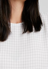 Biały materiał w kratkę z małymi, ciemnymi kwadratami. Dekolt jest okrągły, a materiał wydaje się gładki o miękkiej fakturze.