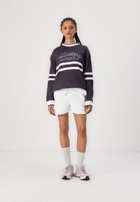 Blusa de manga larga en blanco y negro con logo y rayas, combinada con pantalones cortos blancos. La modelo lleva deportivas claras y calcetines blancos.