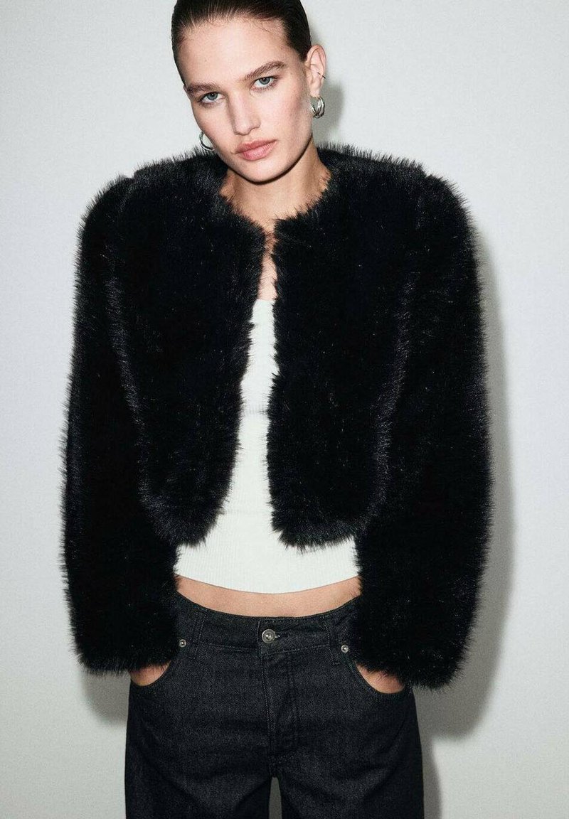 Giacca cropped in faux fur nera con una texture morbida, scollatura tonda e senza abbottonature. Indossata sopra una maglietta bianca e abbinata a jeans denim scuri.