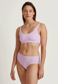 Ženska, oblečena v svetlo vijolično brezšivno modrček in hlačke z visokim pasom, stoji ob enobarvni bež ozadju.