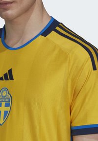 Camiseta de fútbol de tela amarilla con cuello y franjas en los hombros en azul marino; tiene un logo azul y amarillo en el pecho con textura de malla.