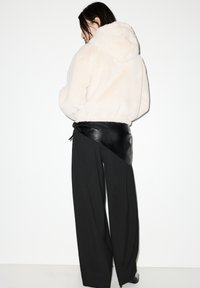 Svijetlosmeđa jakna s kapuljačom od krzna s elastičnim rubom; uparena s visokim crnim hlačama i crnim kožnim remenom za omatanje. Prikazan je stražnji pogled.