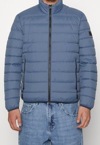Giacca imbottita in morbido tessuto blu con colletto alto, zip frontale e tasche laterali. Presenta cuciture orizzontali e polsini elastici.