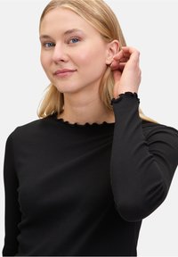 Zwarte langekleding top met geribbeld textuur, featuring een golvende zoom bij de halslijn en manchetten. De pasvorm is dicht op het lichaam.