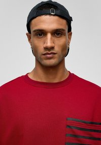 Rood katoenen T-shirt met een gestreepte zwarte zak en een ronde halslijn. Gecombineerd met een zwarte pet. Glad textuur en casual ontwerp.