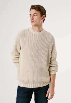 Maglione a coste beige con scollo rotondo e vestibilità morbida. Maniche a cuffia e un'etichetta tessuta sull'orlo aggiungono dettaglio. Indossato con jeans scuri.