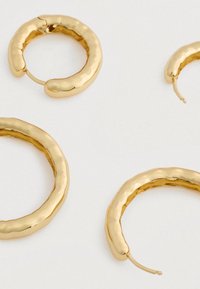 Štyri zlaté textúrované kruhové náušnice rôznych veľkostí rozložené na svetlej ploche, pričom dve majú sponu otvorenú a dve zatvorenú.
