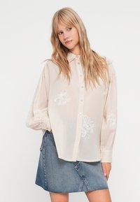 Camisa rosa clara, de botões, com bordado floral, mangas longas e punhos franzidos, combinada com uma saia de denim com uma costura diagonal.