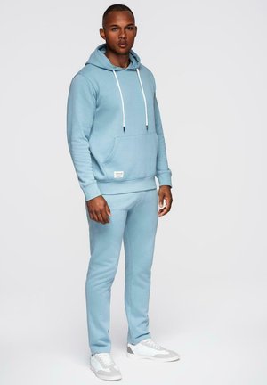 Man staat, draagt een lichtblauwe hoodie en bijpassende joggingbroek met witte en grijze sneakers, tegen een effen achtergrond.