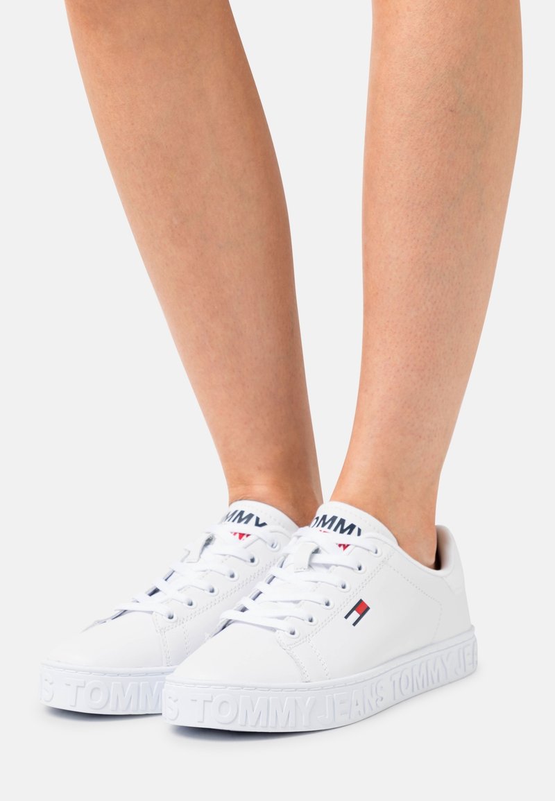 Vita sneakers med en ovandel i läder, platta snören och präglad logotyp på sulan. Röda och blå detaljer nära tungan och sidan.