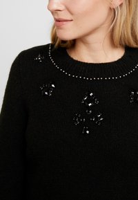 Pull noir en maille orné de perles et de sequins sur le devant, avec un col rond et une texture douce.