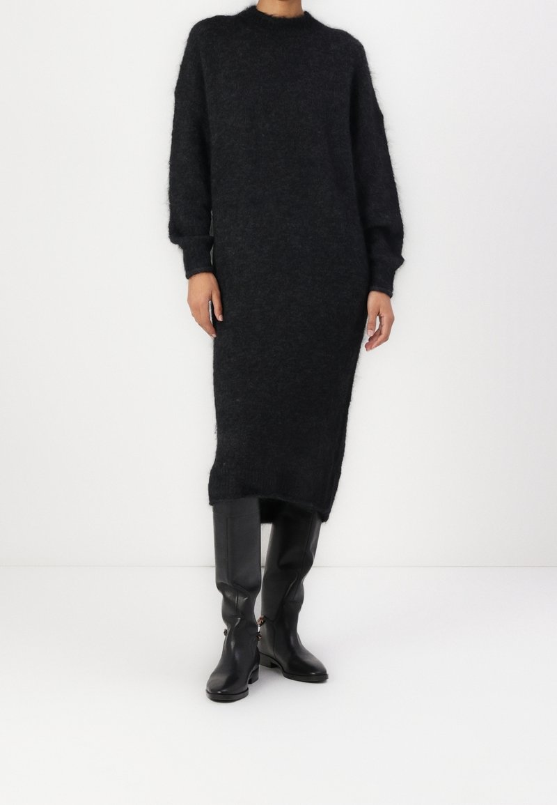 Personne portant une longue robe pull en maille noire et duveteuse ainsi que des bottes en cuir noires montantes jusqu'aux genoux sur un fond blanc uni.