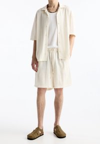 Muž v krémové žebrované košili s krátkými rukávy na zapínání na knoflíky, krátkých šortkách se stahovací šňůrkou, bílém spodním tričku, sandálech s páskem kolem kotníku a náhrdelníku z korálků.