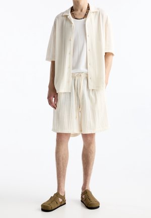 Mies, joka pukeutunut kermaan vivahtavaan ribbineulottuun lyhythihaiseen napitettuun paitaan, siihen sopiviin nyörisomisteisiin shortseihin, valkoiseen aluspaidan alle, nilkkahihallisiin sandaaleihin ja helminauhaan.