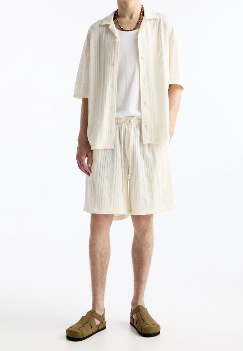 Muž v krémové žebrované košili s krátkými rukávy na zapínání na knoflíky, krátkých šortkách se stahovací šňůrkou, bílém spodním tričku, sandálech s páskem kolem kotníku a náhrdelníku z korálků.