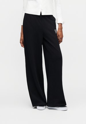 Άτομο που φοράει φαρδιά μαύρη παντελόνα με μικρό λευκό "63" στο μηρό, λευκό πουκάμισο με κουμπιά και μαύρα και λευκά αθλητικά παπούτσια.