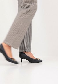 Μαύρα δερμάτινα παπούτσια με ψηλό τακούνι και μυτερή μύτη, με χρυσές λεπτομέρειες, φορεμένα με ριγέ παντελόνια, που αναδεικνύουν έναν κομψό σχεδιασμό.