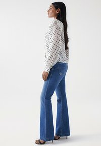Blusa blanca con lunares negros, mangas abullonadas; combinada con jeans de campana azules y sandalias de tacón negro.