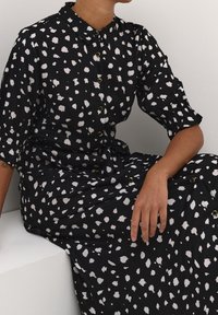 Zwarte jurk met wit spattenpatroon, knoopsluiting aan de voorkant, korte pofmouwen en een losse pasvorm. Gladde stof met gouden knoopdetails.